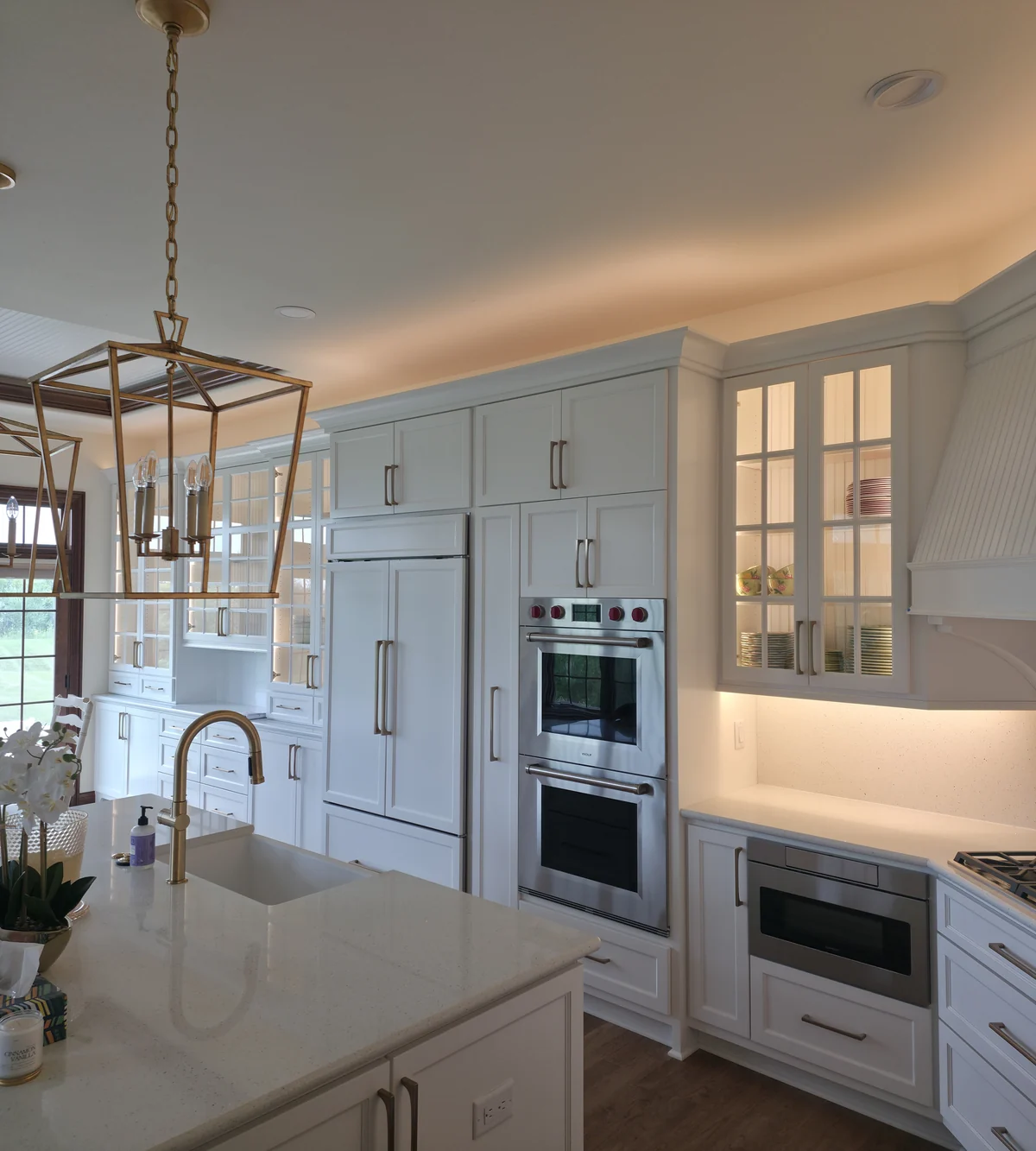 Stainless steel wall ovens and a microwave drawer within white cabinetry, with brass pendants and lit glass cabinets