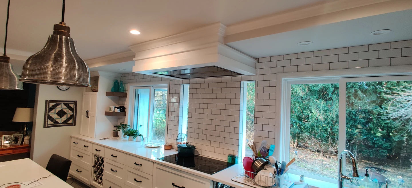 White kitchen with a custom overhead hood, black cooktop, subway tile, and windows overlooking the garden