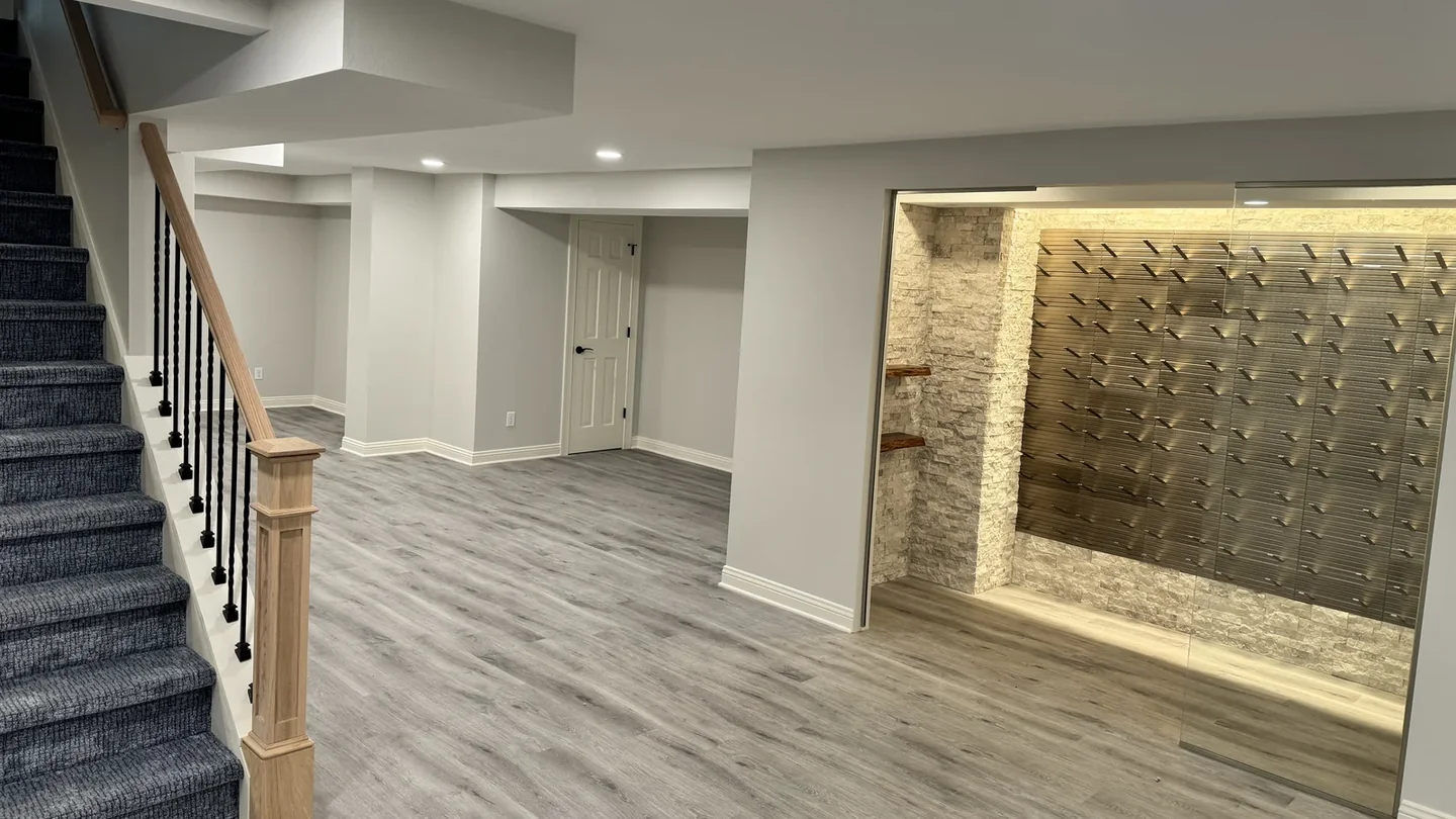 Finished lower level with a wood staircase, gray flooring, and an illuminated glass-enclosed wine display