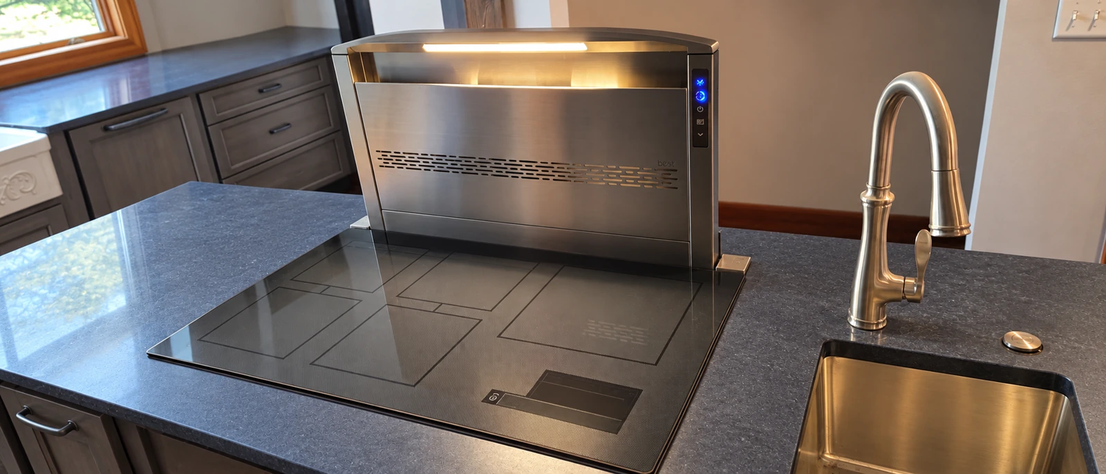 Raised stainless steel downdraft ventilator behind an induction cooktop, beside a brass faucet and kitchen sink