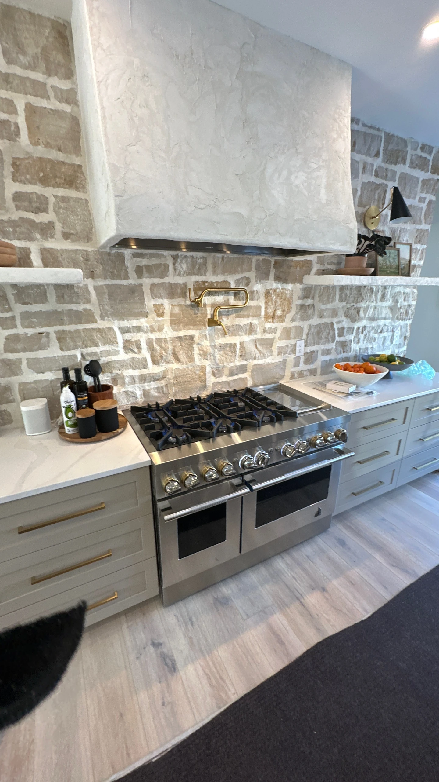 A stainless steel range beneath a sculpted plaster hood, with stone walls, light cabinetry, and brass accents